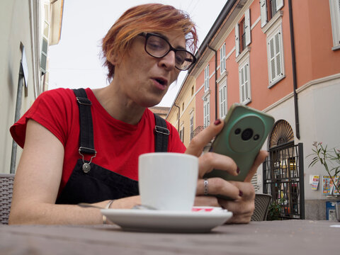 Youthful barista pausing during work shift, wearing red t shirt and dungarees, relaxing at outdoor cafe table while checking smartphone with blurred urban background