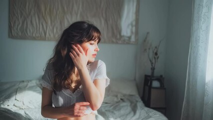 Young woman with long brown hair and bangs wearing a white top and gray shorts sits on a bed with white sheets, posing and looking around in soft sunlight