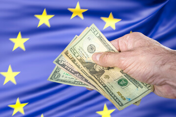 Hand holding some US dollars banknotes in front of a European Union flag, concept of trade war, taxes and tariffs and the 899 Act.