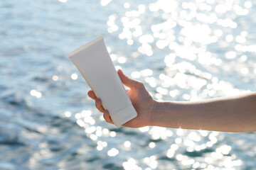 A woman’s hand holds a white cream tube mockup on the sea water background. Skincare concept, sun...
