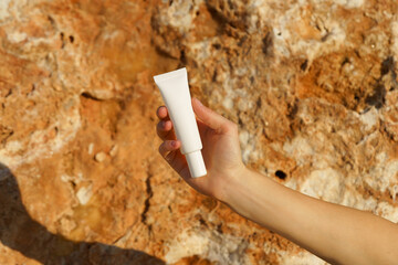 Female hand holding white mockup tube of cream on limestone background near sea on beach. Concept of skin care, moisturizing, protection with Spf, sunscreen. Image for your design
