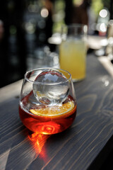 Close-up of a vibrant cocktail with a large ice sphere and orange slice on a wooden table. Natural light enhances the glass, creating a refreshing and inviting scene.