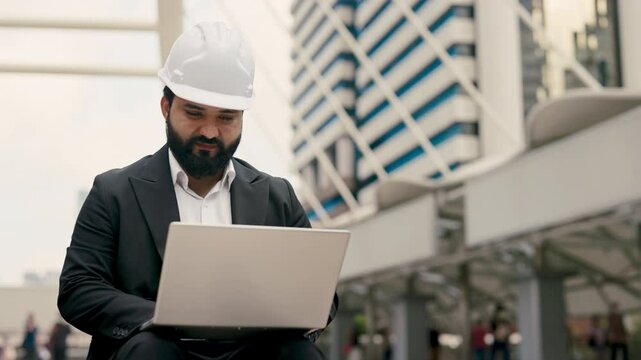 Portrait of focused Indian male manager in helmet and suit working on laptop at office building. Experienced engineer uses notebook computer sitting on parapet of suspension bridge