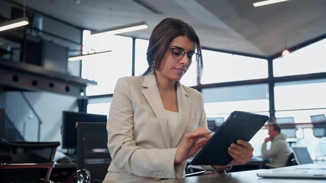 Focused young professional specialist latina hispanic business lady working on tablet pc sitting at desk in modern office. Indian IT manager woman using tab computer device app for work online