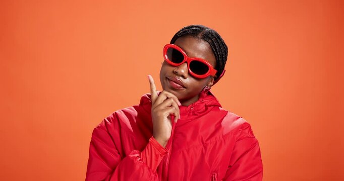 Studio, thinking and woman with sunglasses, fashion and trendy accessory for influencer and serious. Orange background, contemplating and black person with shades, mockup space and colorful jacket