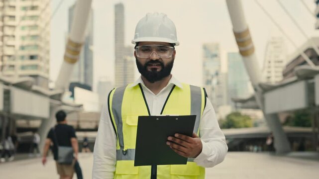Portrait of happy Indian engineer with safety equipment and clipboard at construction site. Bearded technician with architectural plans looks at camera against city bridge