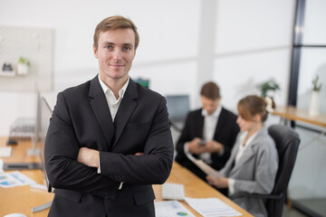 Caucasian businessman with arm crossed gesture in office while colleagues work on laptops in the background