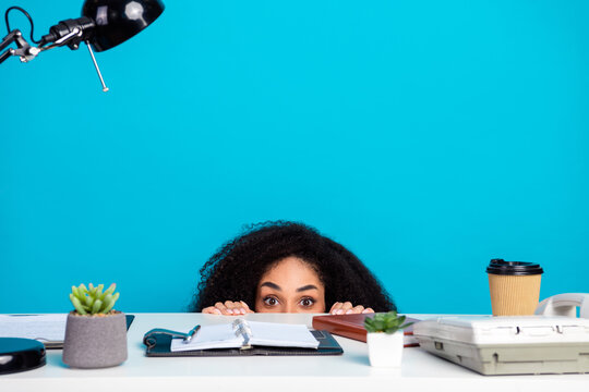 Young woman peeking nervously from behind her workspace desk, surrounded by office items against a bright solid blue background