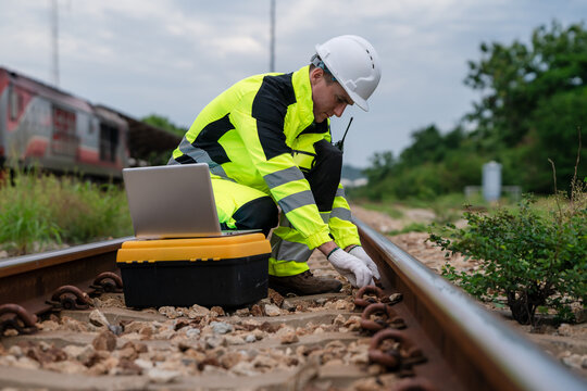 A man in a yellow and black safety suit is working on a train track. He is using a laptop and a cell phone. Inspection and checking of railway tracks, railway maintenance.