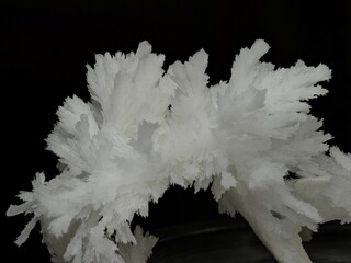 The photo shows a snow-white crystal resembling feathers or snowflakes. Its structure appears fragile and fluffy, with sharp edges highlighted against the black background.