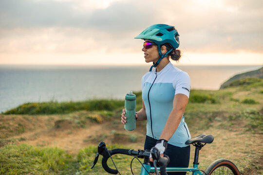 Latin cyclist woman drinking water on gravel bike in front of the sea at sunrise