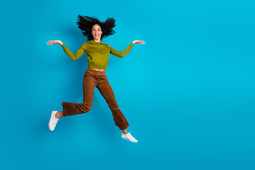 Fototapeta premium Happy young woman jumping joyfully against a blue background wearing casual khaki and brown outfit