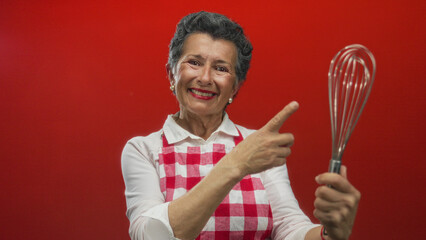 Senior woman with grey hair in apron smiling and gesturing with a whisk against a vibrant red...