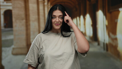Fototapeta premium Young plus size woman making gesture indoors at old university corridor with arches, showcasing confidence and casual style.