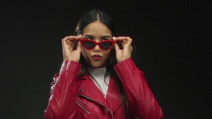 Woman wearing red jacket and sunglasses against a black background exudes confidence and style, her...