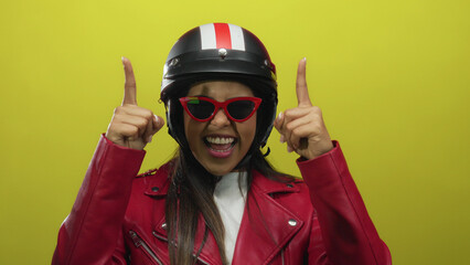 Smiling young woman in red jacket and helmet pointing up with both hands against an isolated yellow background.