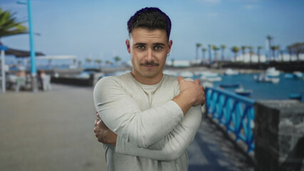 Fototapeta premium Young hispanic man embracing himself on a seaside promenade, with a serene ocean backdrop and clear blue skies, providing a peaceful outdoor setting.