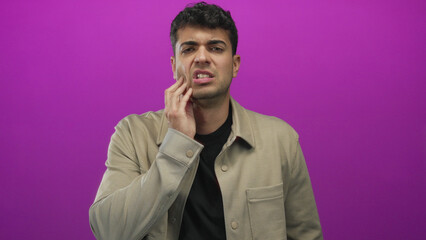 Fototapeta premium Shot captures man cheek touching studio backdrop as young hispanic figure stands against colored purple wall revealing pain and discomfort.