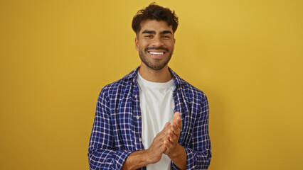Hispanic man claps happily in casual attire against a vibrant yellow background, exuding confidence...