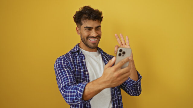 Young hispanic man taking selfie with smartphone against yellow background, smiling in casual attire, capturing a cheerful moment.