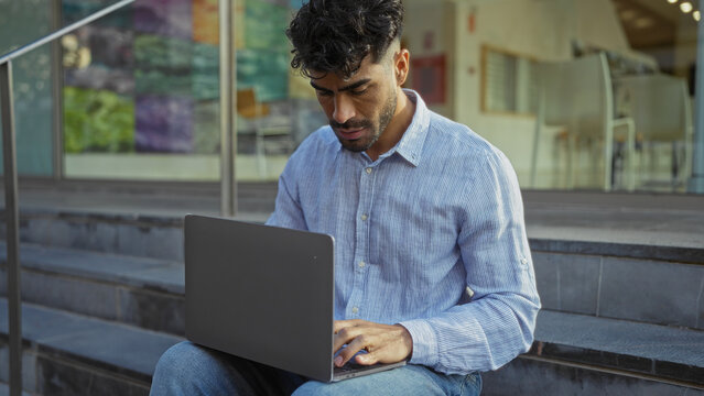 Young man with beard using laptop on urban steps outdoors showcasing focus and technology in a city environment. - Powered by Adobe