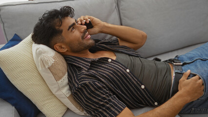 Fototapeta premium Handsome man relaxing indoors on a couch with a phone, casually dressed in a shirt and jeans, exuding charm and comfort in a cozy living room setting.