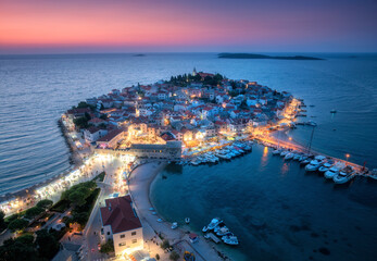 Aeriel view of old town with street lights on island at night. Summer in Primosten, Croatia, Adriatic Sea. Top view of houses, pink sky, water, city lights, boat, yachts, illuminated waterfront
