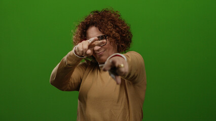Woman making peace gesture in front of green background, middle-aged, curly brunette hair, wearing...