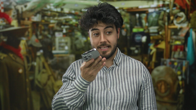 Young hispanic man with beard recording voice message on smartphone in indoor bazar surrounded by eclectic items and wearing striped shirt.