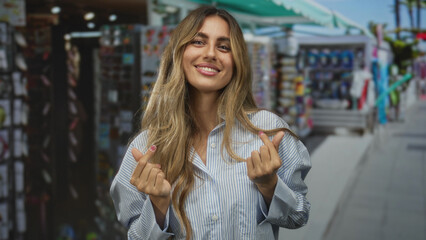 Blonde woman smiling and making finger heart gesture at bustling market stall; love affection joy flirtation.