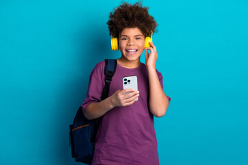 Happy teenager wearing yellow headphones holding a smartphone with a backpack in casual clothing on...