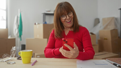 Woman wearing glasses holds smartphone smiling inside new apartment surrounded by moving boxes,...