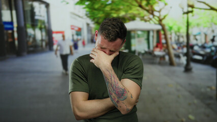 Fototapeta premium Young man with tattoos in an urban street setting expressing strong emotions, wearing a casual green shirt in front of a blurred city background with people walking.