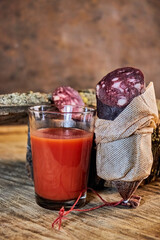 Diagonally sliced ​​​​dry-cured sausage in a traditional fabric casing are served on a wooden surface next to a glass of fresh tomato juice. Serving dishes in a rustic style. 