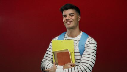 Young hispanic man smiling with backpack and notebooks against a bright red background, wearing a...