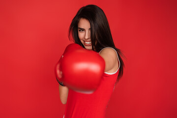 A confident woman poses with boxing gloves, smiling boldly against a vibrant red background,...