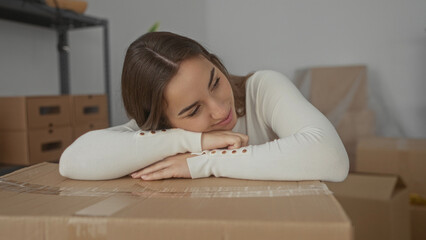 Young woman smiling while leaning on a cardboard box in a new home interior filled with unpacked...