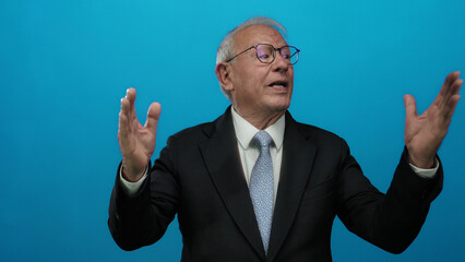 Senior business man gestures passionately against a vibrant blue background wearing a suit and glasses, capturing a moment of expressive communication.