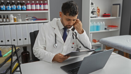 Hispanic male doctor on phone reviewing patient files in clinic office environment featuring...