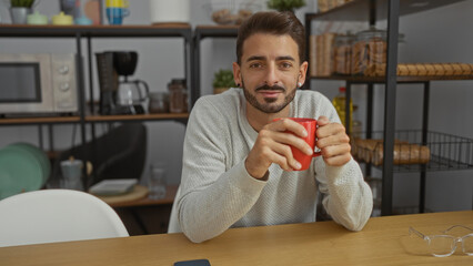 Young hispanic man sitting in office holding red mug with microwave and coffee maker behind on shelf, casual atmosphere conveys professionalism and relaxation