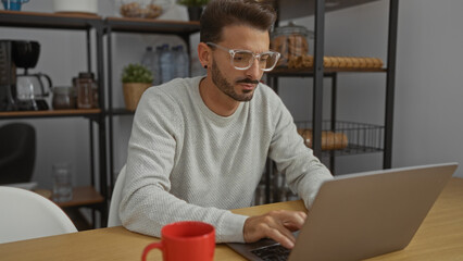 Young man working on laptop in modern office setting with coffee mug on desk and shelves in background portraying focused and productive work environment.