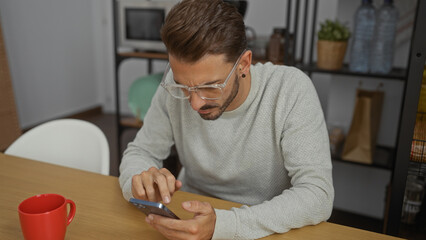 Young man with glasses using smartphone in a modern office environment, focused on his device, with casual attire and a red cup nearby, emphasizing a professional yet relaxed setting.
