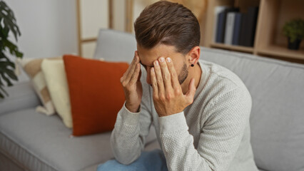 Fototapeta premium Young man sitting on a sofa with hands covering face in a modern apartment living room interior, showing stress and contemplation.