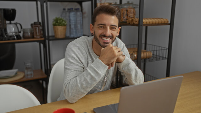 Handsome young man smiling confidently in modern office interior with laptop and shelves in background, creating a friendly workplace atmosphere for creative ideas.