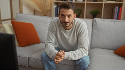 Handsome young man sitting indoors on a cozy sofa in a stylish home living room with modern decor and warm lighting, exuding casual elegance and relaxation.