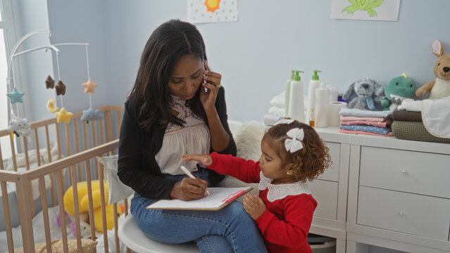 Woman multitasking in bedroom with toddler daughter writing on clipboard near baby cradle surrounded by toys and essentials in cozy home setting