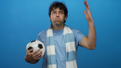 Hispanic man holding soccer ball wearing blue scarf with determined expression against solid blue background depicting sports enthusiasm.