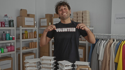Hispanic man smiling proudly in a volunteer shirt stands in a donation room with shelves of...