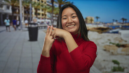 Woman in red sweater making hashtag gesture on sunny seaside promenade with palm trees and sea view, capturing a moment of social media connection outdoor.