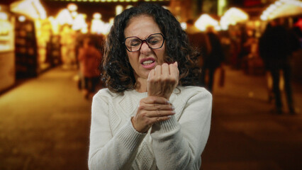 Mature woman with curly hair wearing glasses clutches her wrist in pain on a busy city street at night, surrounded by blurred lights and people.